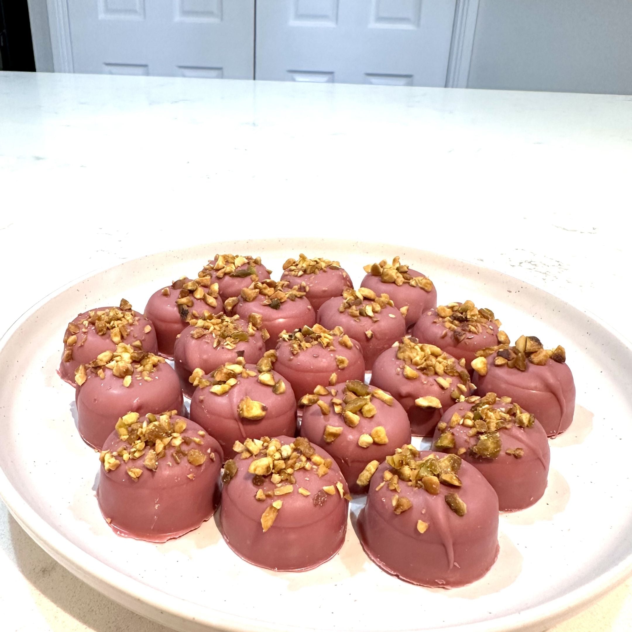 Chocolate On Purpose Ruby Pistachio Truffles with layers of Pashmak (persian ferry floss) and creamy pistachio butter, dipped in pink ruby chocolate, and topped with pistachio, on a white plate. A delicious Valentine’s Day gift idea.