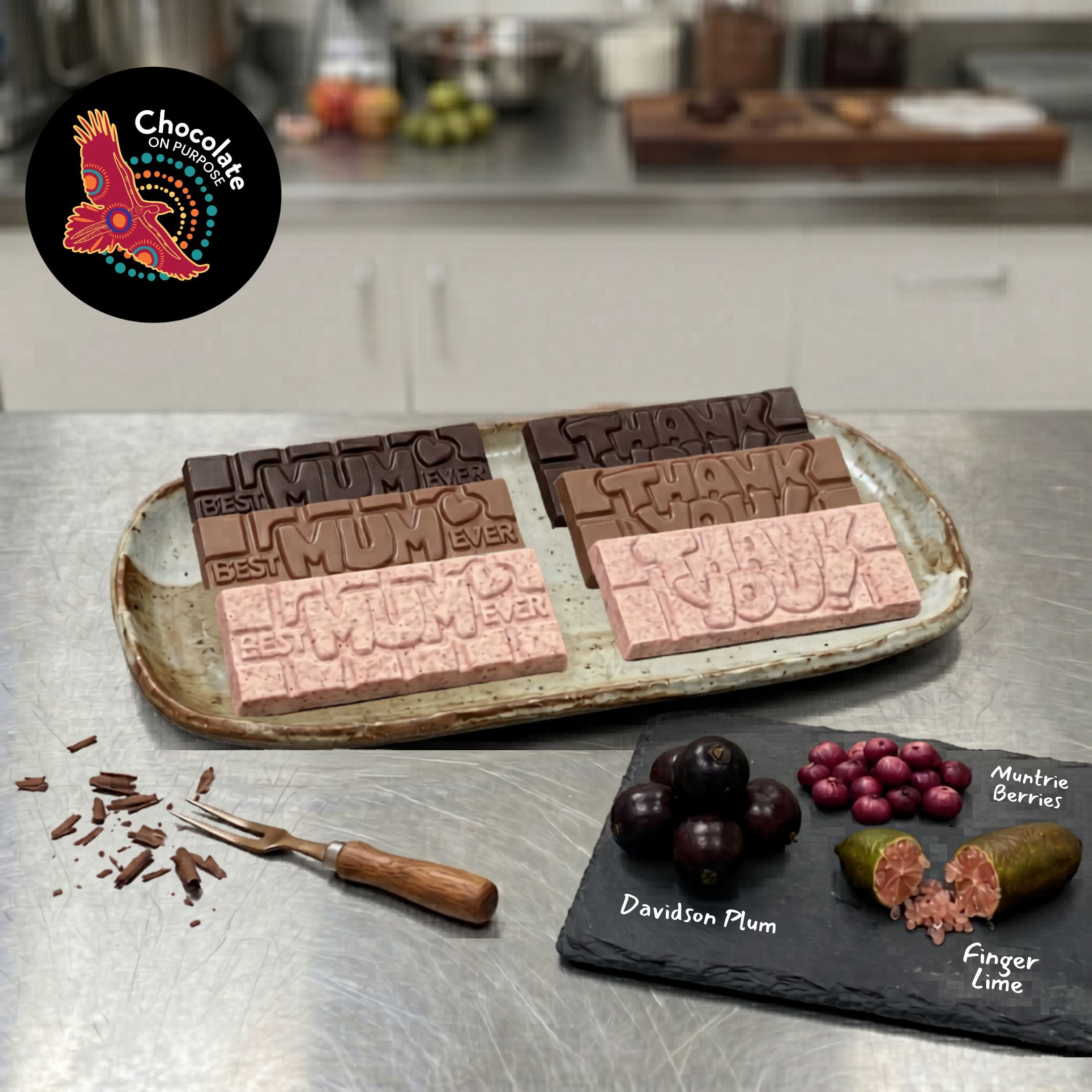 Chocolate On Purpose Mother's Day Bars with embossed 'Best Mum Ever' and 'Thank You Mother's Day' embossed  Bars on a Platter, with Australian native Davidson Plum, Muntrie Berries and Finger Lime fruit on slate plate in front of image. A wooden handled cake fork and grated milk chocolate sit to the left of the slate plate.