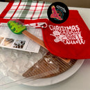 Christmas Chocolate Making Kit flatlay display showing star chocolate mold with clear cavities, red pot holder with "Christmas Calories Don't Count" text and gingerbread man design, red and white checkered tea towel, instruction sheet, piping bag, green Christmas tree plastic spatula, Chocolate On Purpose logo with eagle and dot art on black circle, Indigenous-owned Fair Trade chocolate kit