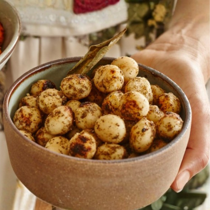 Hand holding a ceramic bowl filled with roasted macadamia nuts, generously coated with Girri Girri (lemon myrtle) and Mourau (mountain pepperberry). The scene highlights the creamy texture of the nuts and the aromatic native botanicals, with a backdrop suggesting a warm, community-focused kitchen. Captures the essence of Aboriginal ingredient knowledge, artisan food, and social impact by Chocolate On Purpose
