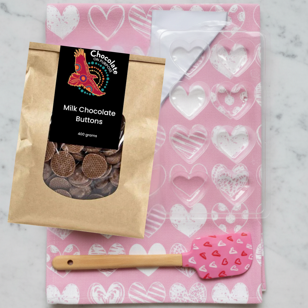 Valentine's Chocolate Making Kit featuring pink tea towel with white heart patterns, clear heart-shaped chocolate mould, milk chocolate buttons, and pink heart-patterned spatula on marble background