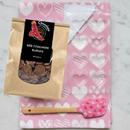 Valentine's Chocolate Making Kit featuring pink tea towel with white heart patterns, clear heart-shaped chocolate mould, milk chocolate buttons, and pink heart-patterned spatula on marble background