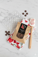 Valentine's Chocolate Making Kit flat lay with heart mould, milk chocolate buttons, red and white tea towel, and red spatula on marble background.