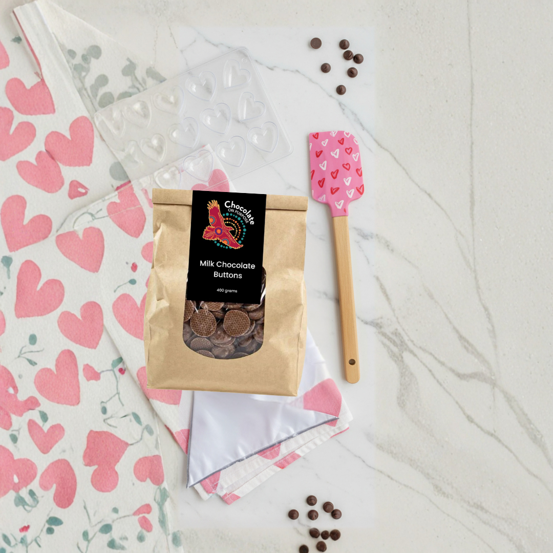 Valentine's Chocolate Making Kit overhead view featuring white tea towel with pink heart pattern, pink heart-patterned spatula, clear heart chocolate mould, milk chocolate buttons, and scattered cocoa nibs on marble