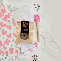 Valentine's Chocolate Making Kit overhead view featuring white tea towel with pink heart pattern, pink heart-patterned spatula, clear heart chocolate mould, milk chocolate buttons, and scattered cocoa nibs on marble