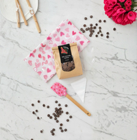 Complete DIY Chocolate Making Kit flat lay with white tea towel featuring assorted pink hearts, pink spatula, heart-shaped chocolate mould, milk chocolate buttons, fresh pink flowers, and chocolate pieces on marble surface