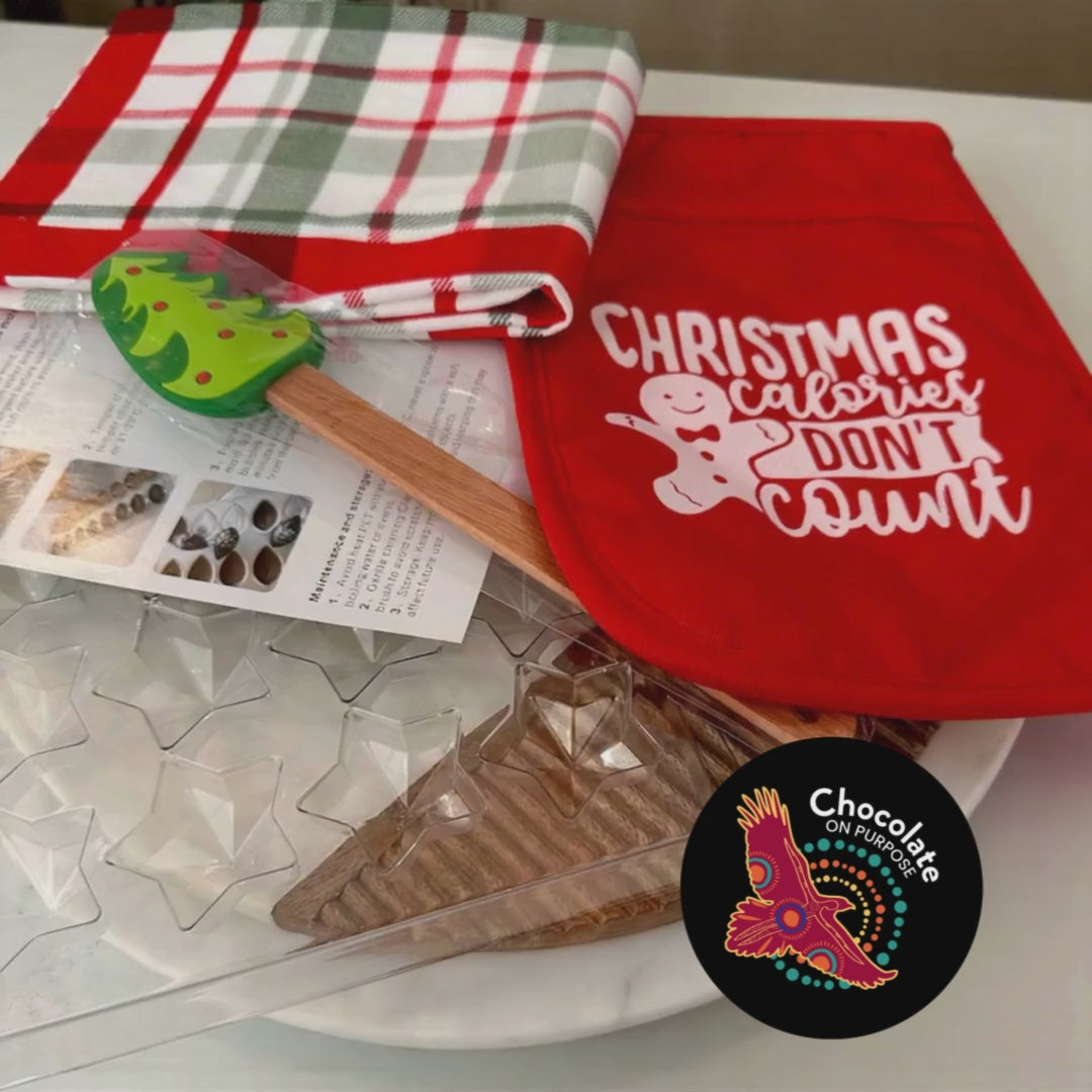 Christmas Chocolate Making Kit flatlay display showing star chocolate mold with clear cavities, red pot holder with "Christmas Calories Don't Count" text and gingerbread man design, red and white checkered tea towel, instruction sheet, piping bag, green Christmas tree plastic spatula, Chocolate On Purpose logo with eagle and dot art on black circle, Indigenous-owned Fair Trade chocolate kit
