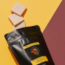 Black bag of White Chocolate with Davidson Plum 3 pieces of chocolate falling out of bag. Yellow Burgundy background
